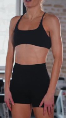 Dikey. Atletik giyinmiş genç bir kadın parlak bir spor salonunda dikiliyor. Spor salonunda kararlı görünen bir kadın. Spor salonundaki antrenmana ara veren zinde bir kadın. Gülümsüyor