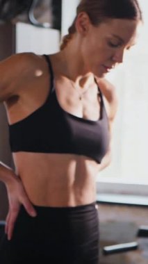 Dikey. Atletik giyinmiş genç bir kadın parlak bir spor salonunda dikiliyor. Spor salonunda kararlı görünen bir kadın. Spor salonundaki antrenmana ara veren zinde bir kadın. Gülümsüyor