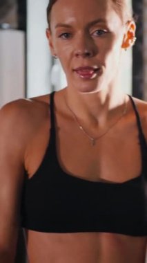 Dikey. Atletik giyinmiş genç bir kadın parlak bir spor salonunda dikiliyor. Spor salonunda kararlı görünen bir kadın. Spor salonundaki antrenmana ara veren zinde bir kadın. Gülümsüyor