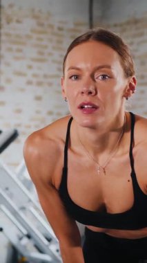 Dikey. Atletik giyinmiş genç bir kadın parlak bir spor salonunda dikiliyor. Spor salonunda kararlı görünen bir kadın. Spor salonundaki antrenmana ara veren zinde bir kadın. Gülümsüyor