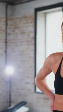 Dikey. Atletik giyinmiş genç bir kadın parlak bir spor salonunda dikiliyor. Spor salonunda kararlı görünen bir kadın. Spor salonundaki antrenmana ara veren zinde bir kadın. Gülümsüyor