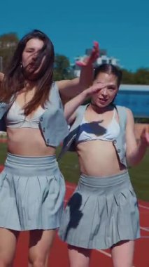 Dikey. Stadyum pistinde neşeyle dans eden iki ateşli kadın. Kısa etekli ponpon kızlar koşu pistinde mutlu bir şekilde dans ediyorlar. İki kız spor pistinde baştan çıkarıcı bir şekilde dans ediyor.