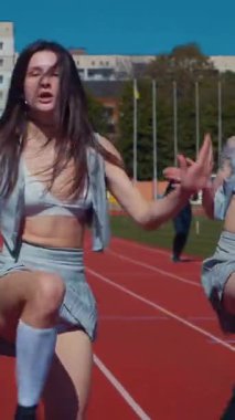 Dikey. Stadyum pistinde neşeyle dans eden iki ateşli kadın. Kısa etekli ponpon kızlar koşu pistinde mutlu bir şekilde dans ediyorlar. İki kız spor pistinde baştan çıkarıcı bir şekilde dans ediyor.