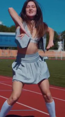 Dikey. Stadyum pistinde neşeyle dans eden iki ateşli kadın. Kısa etekli ponpon kızlar koşu pistinde mutlu bir şekilde dans ediyorlar. İki kız spor pistinde baştan çıkarıcı bir şekilde dans ediyor.