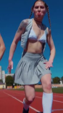 Dikey. Stadyum pistinde neşeyle dans eden iki ateşli kadın. Kısa etekli ponpon kızlar koşu pistinde mutlu bir şekilde dans ediyorlar. İki kız spor pistinde baştan çıkarıcı bir şekilde dans ediyor.