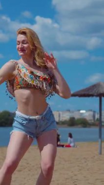 Dikey. Bachata, salsa ve kizomba yapan bir kadın. Sahilde, mavi gökyüzünün arka planında. Gülümseyen kadın çıplak ayakla poz verir ve sıcak kumda baştan çıkarıcı bir şekilde dans eder. Plaj sahnesinde kızın keyfi yerinde. Şortlu ve gömlekli güzel bayan. 