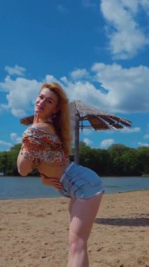 Dikey. Bachata, salsa ve kizomba yapan bir kadın. Sahilde, mavi gökyüzünün arka planında. Gülümseyen kadın çıplak ayakla poz verir ve sıcak kumda baştan çıkarıcı bir şekilde dans eder. Plaj sahnesinde kızın keyfi yerinde. Şortlu ve gömlekli güzel bayan. 