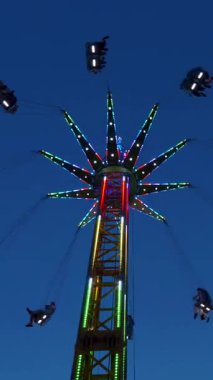 Dikey. Gece gökyüzüne karşı parlayan ışıklarla süslenmiş canlı bir eğlence gezisi. Luna Parkı. Parlak yeşil ışıklar lunaparktaki dönen bir atlıkarıncanın üzerinde dans ediyor. Ekstrem atlıkarınca neşeyle dönüyor, büyüleyici yeşil ışıklarla aydınlatılıyor