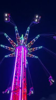 Dikey. Gece gökyüzüne karşı parlayan ışıklarla süslenmiş canlı bir eğlence gezisi. Luna Parkı. Parlak yeşil ışıklar lunaparktaki dönen bir atlıkarıncanın üzerinde dans ediyor. Ekstrem atlıkarınca neşeyle dönüyor, büyüleyici yeşil ışıklarla aydınlatılıyor