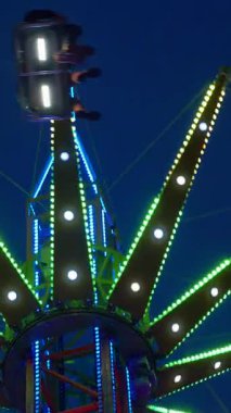 Dikey. Gece gökyüzüne karşı parlayan ışıklarla süslenmiş canlı bir eğlence gezisi. Luna Parkı. Parlak yeşil ışıklar lunaparktaki dönen bir atlıkarıncanın üzerinde dans ediyor. Ekstrem atlıkarınca neşeyle dönüyor, büyüleyici yeşil ışıklarla aydınlatılıyor