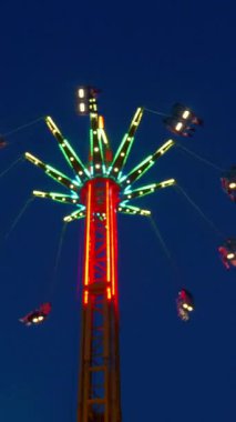 Dikey. Gece gökyüzüne karşı parlayan ışıklarla süslenmiş canlı bir eğlence gezisi. Luna Parkı. Parlak yeşil ışıklar lunaparktaki dönen bir atlıkarıncanın üzerinde dans ediyor. Ekstrem atlıkarınca neşeyle dönüyor, büyüleyici yeşil ışıklarla aydınlatılıyor