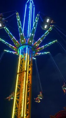 Dikey. Gece gökyüzüne karşı parlayan ışıklarla süslenmiş canlı bir eğlence gezisi. Luna Parkı. Parlak yeşil ışıklar lunaparktaki dönen bir atlıkarıncanın üzerinde dans ediyor. Ekstrem atlıkarınca neşeyle dönüyor, büyüleyici yeşil ışıklarla aydınlatılıyor