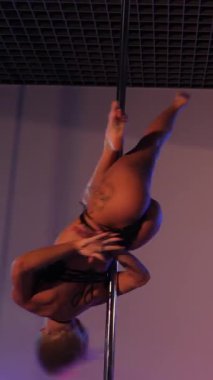 Dikey. Tangalı güzel bir kadın direk dansı yapıyor. Direğin üstünde erotik dans. Gece kulübü. Yüksek topuklu ayakkabı, kutup dansı için hassas bir dokunuş, büyüleyici bir performans. Büyüleyici güç ve esneklik gösterisi.