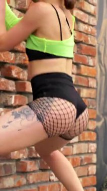 Dikey. Sokakta siyah bir duvarın yanında dans eden kız. Erotik dans eden kadın. Genç kız enerjik bir şekilde seksi bir dans ediyor. Çıplak kalçalarla dans eden bir kız baştan çıkarıcı bir dansla.