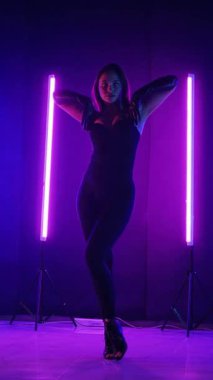 Dikey. Siyah elbiseli, neon ışıklı, baştan çıkarıcı dans performansı. Yüksek topuklu. Güzel kız erotik olarak kameraya poz veriyor. Parlak neon ışıklı dans pisti