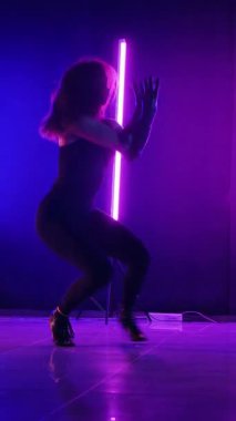 Dikey. Siyah elbiseli, neon ışıklı, baştan çıkarıcı dans performansı. Yüksek topuklu. Güzel kız erotik olarak kameraya poz veriyor. Parlak neon ışıklı dans pisti