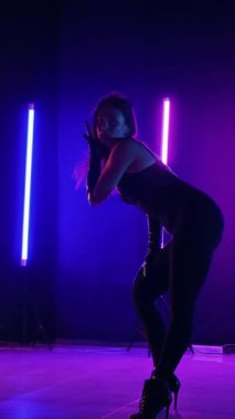 Dikey. Siyah elbiseli, neon ışıklı, baştan çıkarıcı dans performansı. Yüksek topuklu. Güzel kız erotik olarak kameraya poz veriyor. Parlak neon ışıklı dans pisti