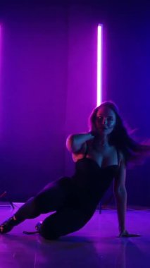 Dikey. Siyah elbiseli, neon ışıklı, baştan çıkarıcı dans performansı. Yüksek topuklu. Güzel kız erotik olarak kameraya poz veriyor. Parlak neon ışıklı dans pisti