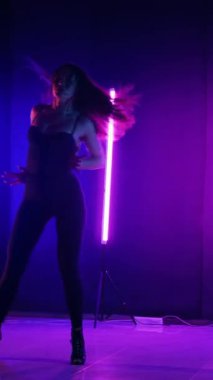 Dikey. Siyah elbiseli, neon ışıklı, baştan çıkarıcı dans performansı. Yüksek topuklu. Güzel kız erotik olarak kameraya poz veriyor. Parlak neon ışıklı dans pisti
