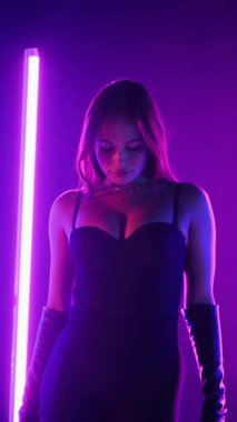 Dikey. Siyah elbiseli, neon ışıklı, baştan çıkarıcı dans performansı. Yüksek topuklu. Güzel kız erotik olarak kameraya poz veriyor. Parlak neon ışıklı dans pisti