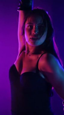 Dikey. Siyah elbiseli, neon ışıklı, baştan çıkarıcı dans performansı. Yüksek topuklu. Güzel kız erotik olarak kameraya poz veriyor. Parlak neon ışıklı dans pisti