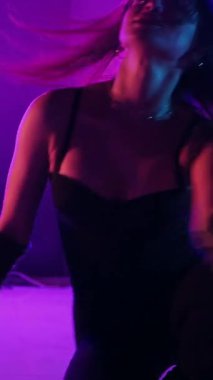 Dikey. Siyah elbiseli, neon ışıklı, baştan çıkarıcı dans performansı. Yüksek topuklu. Güzel kız erotik olarak kameraya poz veriyor. Parlak neon ışıklı dans pisti