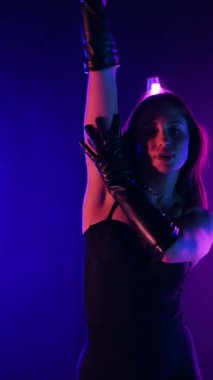 Dikey. Siyah elbiseli, neon ışıklı, baştan çıkarıcı dans performansı. Yüksek topuklu. Güzel kız erotik olarak kameraya poz veriyor. Parlak neon ışıklı dans pisti