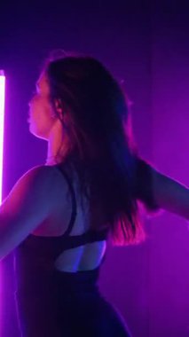 Dikey. Siyah elbiseli, neon ışıklı, baştan çıkarıcı dans performansı. Yüksek topuklu. Güzel kız erotik olarak kameraya poz veriyor. Parlak neon ışıklı dans pisti