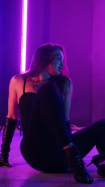 Dikey. Siyah elbiseli, neon ışıklı, baştan çıkarıcı dans performansı. Yüksek topuklu. Güzel kız erotik olarak kameraya poz veriyor. Parlak neon ışıklı dans pisti
