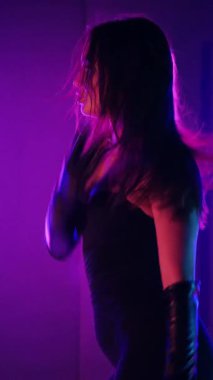 Dikey. Siyah elbiseli, neon ışıklı, baştan çıkarıcı dans performansı. Yüksek topuklu. Güzel kız erotik olarak kameraya poz veriyor. Parlak neon ışıklı dans pisti