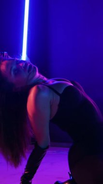 Dikey. Siyah elbiseli, neon ışıklı, baştan çıkarıcı dans performansı. Yüksek topuklu. Güzel kız erotik olarak kameraya poz veriyor. Parlak neon ışıklı dans pisti