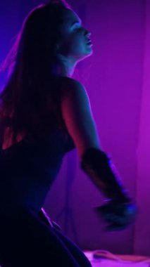 Dikey. Siyah elbiseli, neon ışıklı, baştan çıkarıcı dans performansı. Yüksek topuklu. Güzel kız erotik olarak kameraya poz veriyor. Parlak neon ışıklı dans pisti