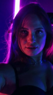 Dikey. Siyah elbiseli, neon ışıklı, baştan çıkarıcı dans performansı. Yüksek topuklu. Güzel kız erotik olarak kameraya poz veriyor. Parlak neon ışıklı dans pisti