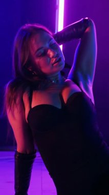 Dikey. Siyah elbiseli, neon ışıklı, baştan çıkarıcı dans performansı. Yüksek topuklu. Güzel kız erotik olarak kameraya poz veriyor. Parlak neon ışıklı dans pisti