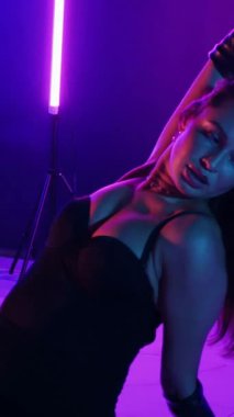 Dikey. Siyah elbiseli, neon ışıklı, baştan çıkarıcı dans performansı. Yüksek topuklu. Güzel kız erotik olarak kameraya poz veriyor. Parlak neon ışıklı dans pisti