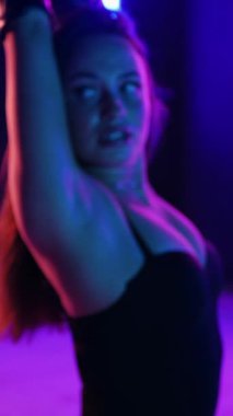 Dikey. Siyah elbiseli, neon ışıklı, baştan çıkarıcı dans performansı. Yüksek topuklu. Güzel kız erotik olarak kameraya poz veriyor. Parlak neon ışıklı dans pisti