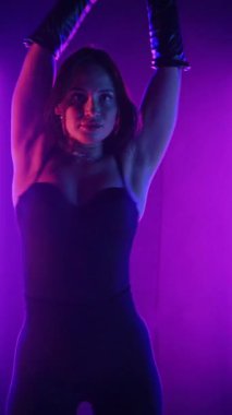 Dikey. Siyah elbiseli, neon ışıklı, baştan çıkarıcı dans performansı. Yüksek topuklu. Güzel kız erotik olarak kameraya poz veriyor. Parlak neon ışıklı dans pisti