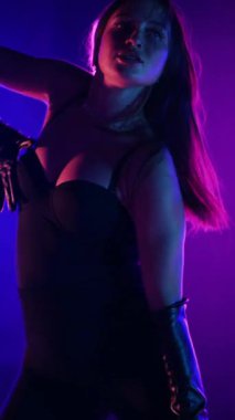 Dikey. Siyah elbiseli, neon ışıklı, baştan çıkarıcı dans performansı. Yüksek topuklu. Güzel kız erotik olarak kameraya poz veriyor. Parlak neon ışıklı dans pisti