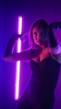 Dikey. Siyah elbiseli, neon ışıklı, baştan çıkarıcı dans performansı. Yüksek topuklu. Güzel kız erotik olarak kameraya poz veriyor. Parlak neon ışıklı dans pisti