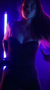 Dikey. Siyah elbiseli, neon ışıklı, baştan çıkarıcı dans performansı. Yüksek topuklu. Güzel kız erotik olarak kameraya poz veriyor. Parlak neon ışıklı dans pisti
