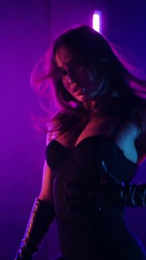 Dikey. Siyah elbiseli, neon ışıklı, baştan çıkarıcı dans performansı. Yüksek topuklu. Güzel kız erotik olarak kameraya poz veriyor. Parlak neon ışıklı dans pisti