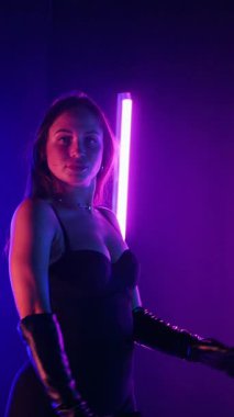 Dikey. Siyah elbiseli, neon ışıklı, baştan çıkarıcı dans performansı. Yüksek topuklu. Güzel kız erotik olarak kameraya poz veriyor. Parlak neon ışıklı dans pisti