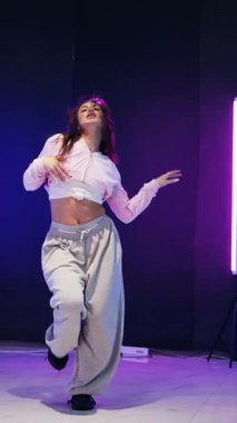 Dikey. Enerjik kız caz funk, hip hop, canlı neon fon ile dans ediyor. Genç kadın neon ışığı altında zarifçe hareket ediyor. Renkli neon ışıkların önünde dans eden kız. Neon ışıkları kızın neşesini aydınlatır.