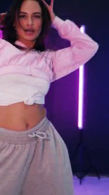 Dikey. Enerjik kız caz funk, hip hop, canlı neon fon ile dans ediyor. Genç kadın neon ışığı altında zarifçe hareket ediyor. Renkli neon ışıkların önünde dans eden kız. Neon ışıkları kızın neşesini aydınlatır.