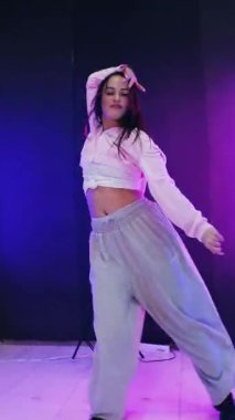 Dikey. Enerjik kız caz funk, hip hop, canlı neon fon ile dans ediyor. Genç kadın neon ışığı altında zarifçe hareket ediyor. Renkli neon ışıkların önünde dans eden kız. Neon ışıkları kızın neşesini aydınlatır.