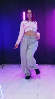 Dikey. Enerjik kız caz funk, hip hop, canlı neon fon ile dans ediyor. Genç kadın neon ışığı altında zarifçe hareket ediyor. Renkli neon ışıkların önünde dans eden kız. Neon ışıkları kızın neşesini aydınlatır.