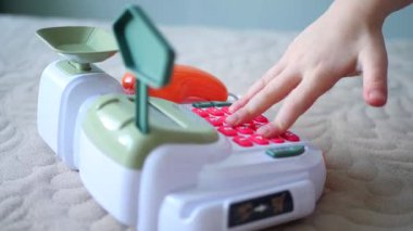 Bir çocuğun elleri oyuncak kasasının renkli tuşlarına basıyor. Minyatür bir kasadaki düğmelere titizlikle basan bir çocuğun parmak görüntüsü. Hesap makinesi, ölçek, banka terminali olan çocuk kasası. Oyuncak para ve bozukluklar