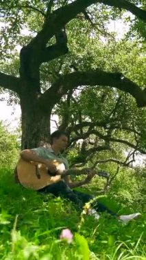 Dikey. Yapraklı bir gölgelik altında akustik gitar çalan bir adam, benekli güneş ışığıyla yıkanmış. nazik tıngırtı, yemyeşil, ve huzurlu bir melodi. Akustik gitar müziği orman havasını doldurur, bir adamın parmakları tellerde dans eder.. 