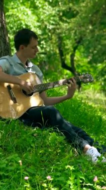 Dikey. Yapraklı bir gölgelik altında akustik gitar çalan bir adam, benekli güneş ışığıyla yıkanmış. nazik tıngırtı, yemyeşil, ve huzurlu bir melodi. Akustik gitar müziği orman havasını doldurur, bir adamın parmakları tellerde dans eder.. 