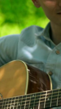 Dikey. Yapraklı bir gölgelik altında akustik gitar çalan bir adam, benekli güneş ışığıyla yıkanmış. nazik tıngırtı, yemyeşil, ve huzurlu bir melodi. Akustik gitar müziği orman havasını doldurur, bir adamın parmakları tellerde dans eder.. 
